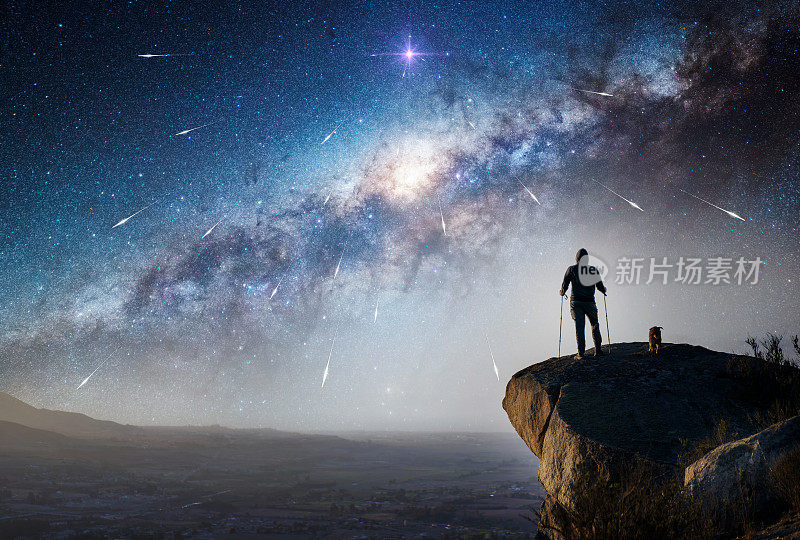 一个徒步旅行者和他的狗在山顶凝视银河和地平线上的流星的剪影