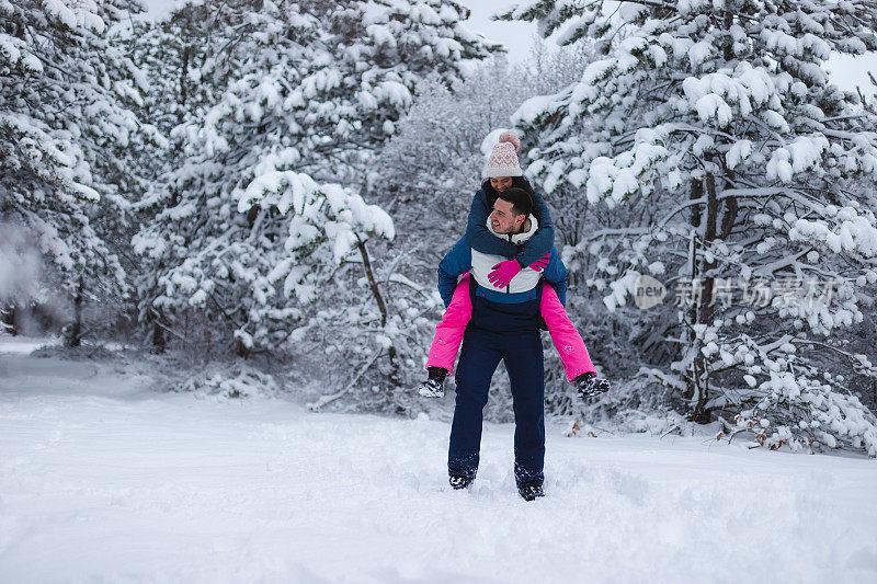 年轻的男朋友背着他的女朋友在雪地里玩，周围是雪树