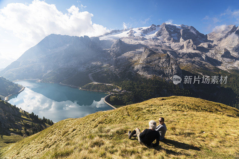 在意大利北部Dolomites的Fedaia山口到Pordoi山口的小路上休息的老年人夫妇正在欣赏美丽的Fedaia湖和马尔莫拉达山的景色。