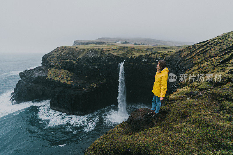 穿着黄色雨衣的女人在看风景Múlafossur瀑布掉进海里在法罗群岛