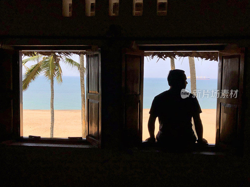 在印度南部果阿附近的喀拉拉邦，度假男子的剪影坐在窗前，在茅草海滩房子的木制百叶窗打开，放松和看远处的热带海滩景色，太阳，大海，沙子和椰子树的照片