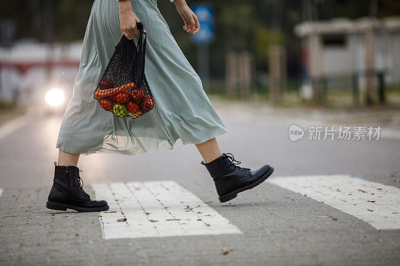 穿着飘逸裙子的年轻女子提着装着食品杂货的网袋过马路