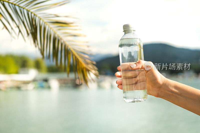 女性手拿一个装着水的塑料瓶，旁边是一棵棕榈树。