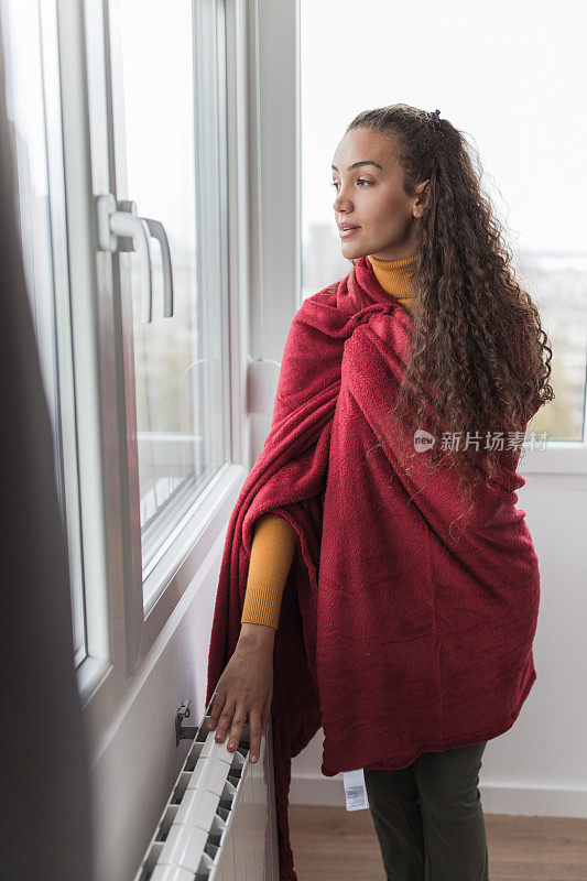 裹着毯子的严肃年轻女子站在窗前，触摸着冰冷的散热器