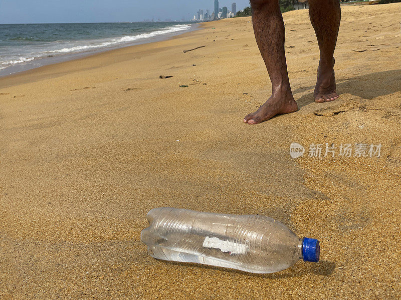 特写图像，空的透明塑料水瓶，蓝色盖子被冲到斯里兰卡热带海滩的金色沙滩上，旁边是无法辨认的人，从拉维尼亚山海滩上看到科伦坡市摩天大楼的海岸线，复制空间