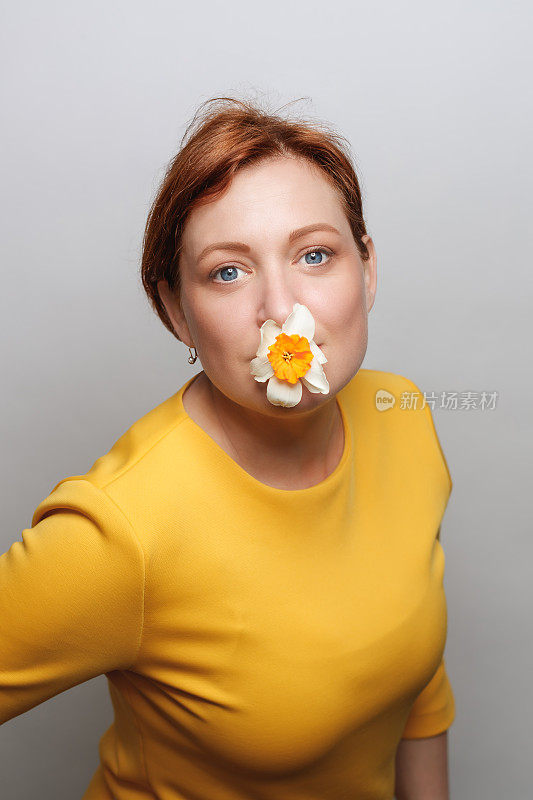 有趣的肖像中年妇女在黄色上衣拿着水仙花在嘴里，看着相机