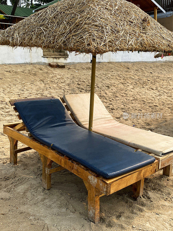 海滩度假的特写图像，一对木制，蓝色和奶油软垫的太阳躺椅床，在稻草遮阳伞下在沙滩上日光浴，擦拭干净的靠垫，果阿，印度