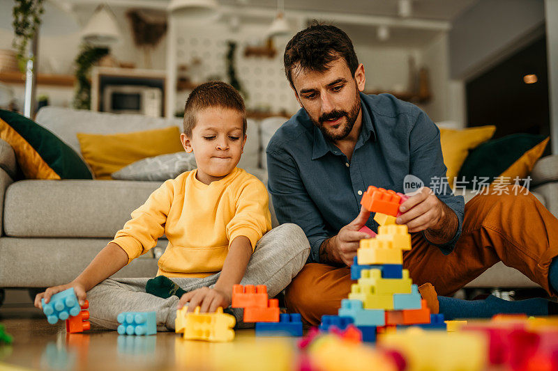 一位充满爱心的单身父亲带着可爱的儿子一起在温暖的地板上玩耍，一位快乐的家庭父亲和小男孩一起用五颜六色的积木搭起了一座建筑塔。
