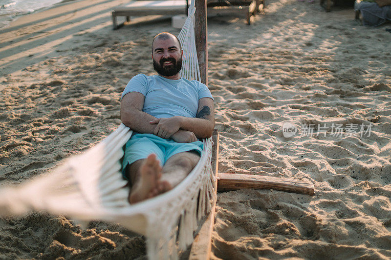 阳光明媚的一天，一个满脸笑容的大胡子男子躺在沙滩上的吊床上休息