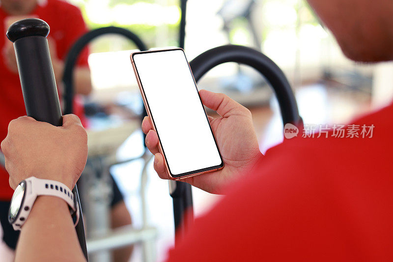 一位亚洲中年男子在健身房锻炼时，花了一点时间用智能手机上的健身追踪应用检查自己的脉搏和训练表现。智能手机上的空白屏幕用于设计模型。