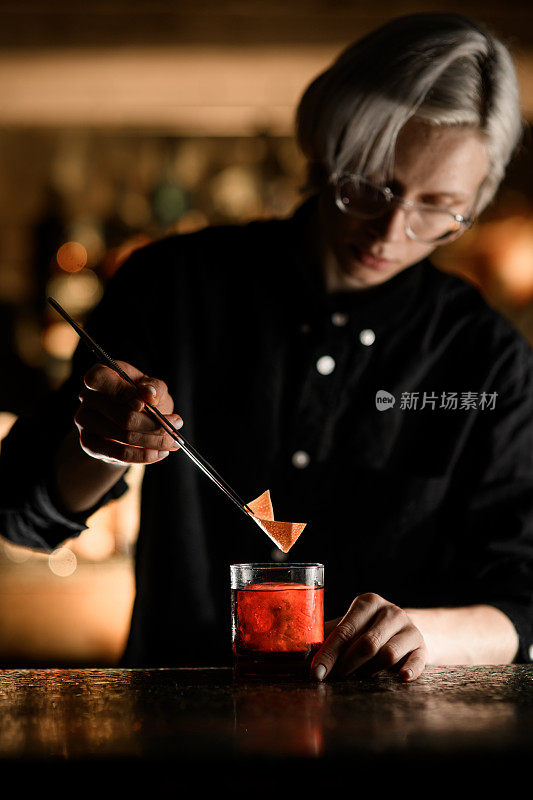 女调酒师用镊子从装有深红色冰鸡尾酒的玻璃杯中取出一块橘子皮