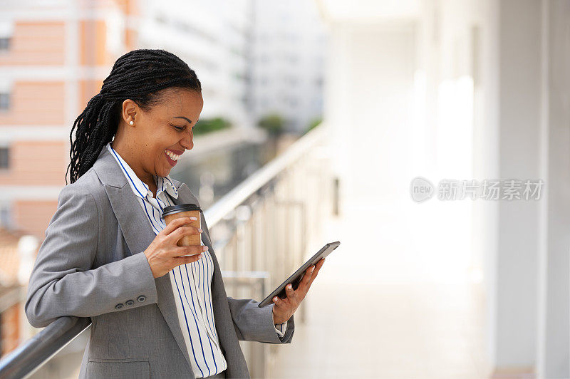 一位非裔美国女商人站在办公楼的阳台上，休息一下，喝着咖啡，玩着一台小型平板电脑