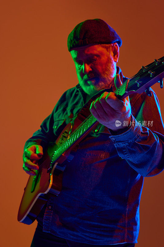 有胡子的吉他手专注地调整电吉他在彩色霓虹灯对橙色工作室背景。