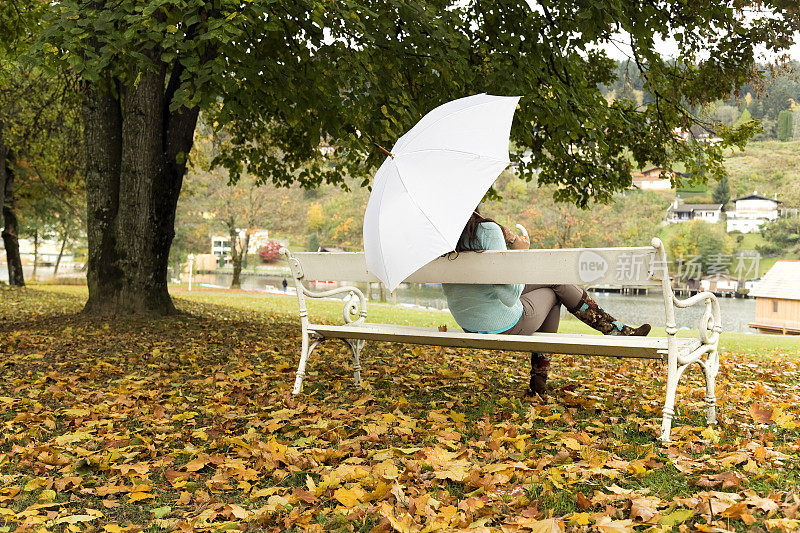 秋天里，拿着白伞的女人坐在公园的长椅上