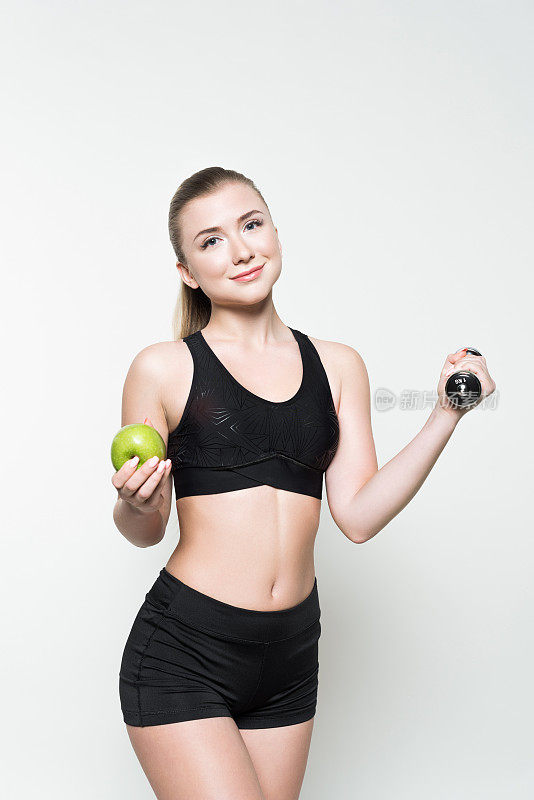 年轻女子在运动上衣举着哑铃和苹果孤立在白色