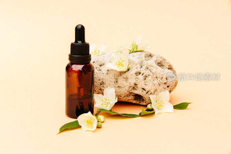 漂亮的茶油组合。玻璃化妆品瓶立在一块石头上，米色的背景上有茉莉花，明亮的阳光。天然化妆品概念，天然精油。