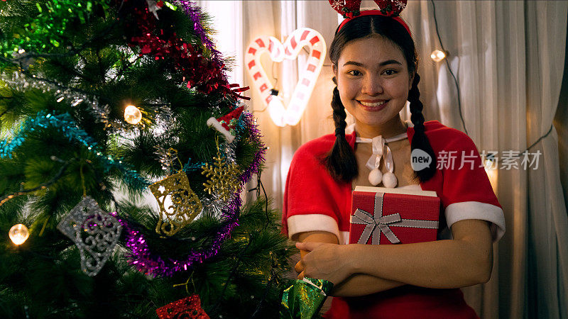 亚洲妇女穿着圣诞老人角色扮演服装，她装饰圣诞树在客厅靠窗的房子里面。