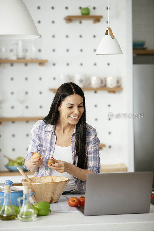 快乐的女人一边在厨房里准备午餐，一边用手提电脑上网。库存图片