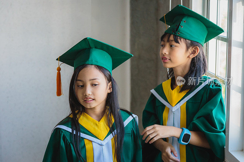 肖像可爱的两个亚洲小女孩戴着毕业帽和长袍站在教室在学校，小学和学生的概念