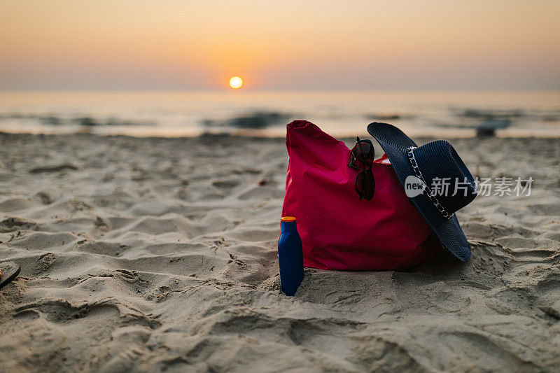 一个海滩袋的特写，帽子，防晒霜和太阳眼镜在沙滩上与日落在海上的背景