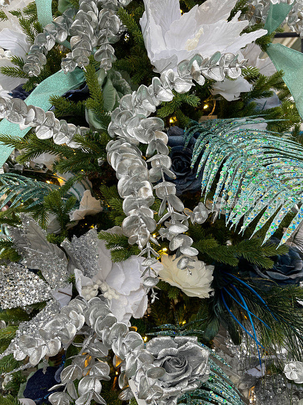 形象的人造，银色的桉树树叶，绿色的亮片树叶，白色和蓝色的玫瑰花头装饰品挂在人造圣诞树的树枝上，绿色的丝带，云杉针，模糊的白色神仙灯的背景