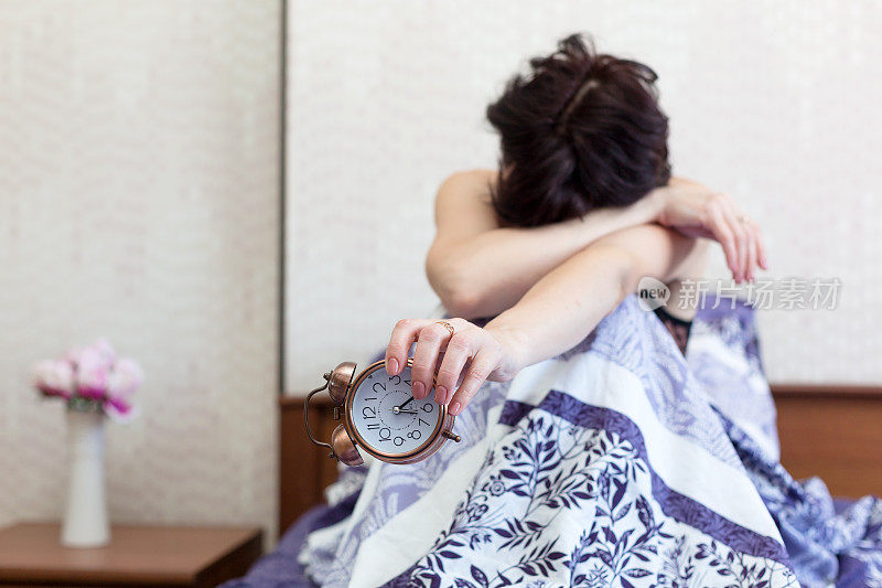 一个昏昏欲睡的女人坐在床上，脸埋在膝盖里，手里拿着一个闹钟。不入睡的概念，重醒
