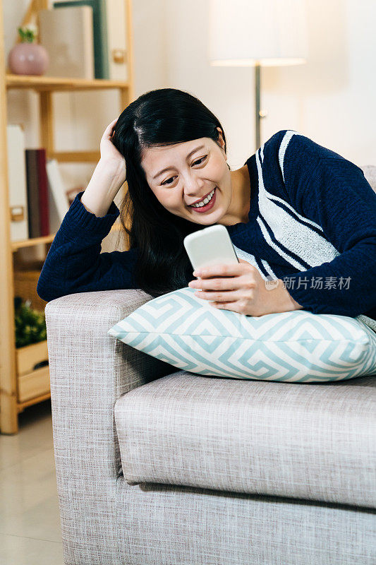 垂直拍摄华丽的女士支撑在沙发上的头享受浏览社交媒体饲料在她的手机休闲。技术、生活方式、室内娱乐理念。