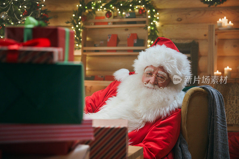 笑容可掬的圣诞老人从一堆礼品盒后面看着我们，他半转着身子坐在堆满圣诞装饰的书房里的扶手椅上