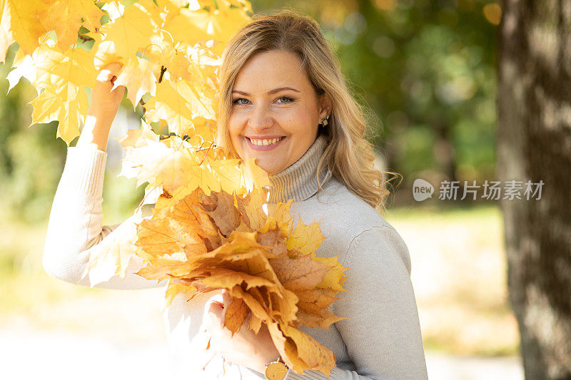 美丽微笑的中年妇女，卷曲的金发，卷领毛衣，捧着一束枫叶和树枝。