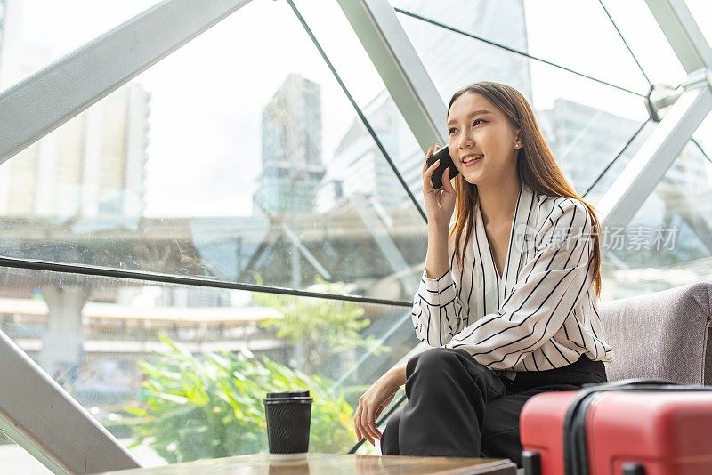 美丽的年轻亚洲女性商务独自女旅行者在机场候机楼的咖啡馆里等待起飞时用智能手机打电话