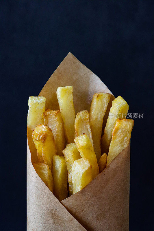 图像的防油羊皮纸包鲜煮的脆的自制炸薯条，包着一堆金黄色的薯片零食，黑色背景，重点在前景