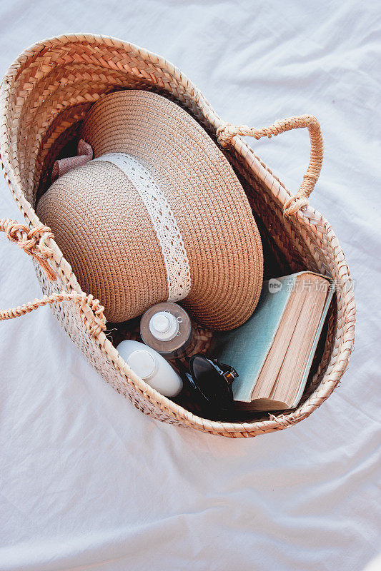 垂直拍摄一个草编袋子，里面有草帽、防晒霜、一本书和太阳镜