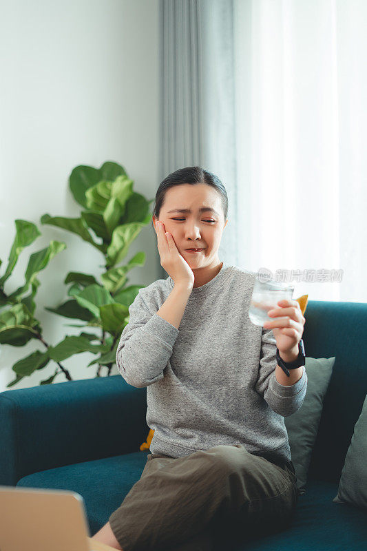 亚洲女性坐在客厅的沙发上，喝了凉水，摸了摸脸颊，就会感到牙齿敏感。