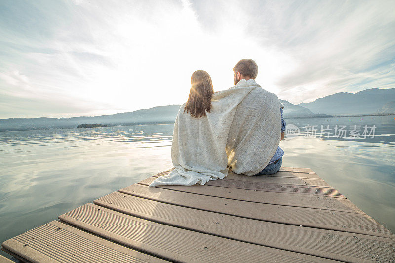一对年轻夫妇裹着毯子在湖边码头上休息