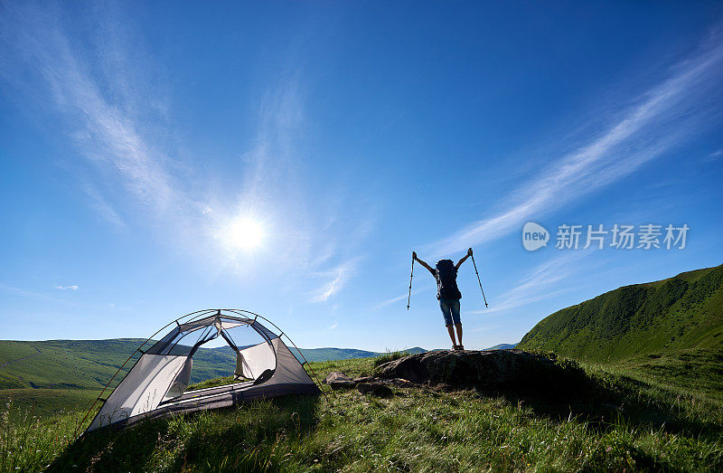 女游客背景站在山顶，顶着帐篷附近的蓝天、阳光和云彩，手拿登山杖，在空中举起双手，享受山中阳光明媚的早晨
