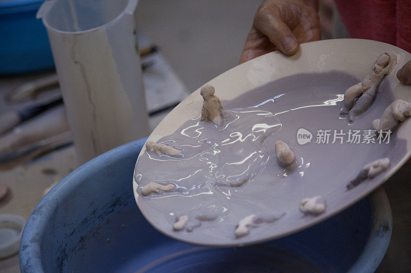 艺术家用手将未完成的陶瓷作品浸在蓝釉中，艺术盘中有小人