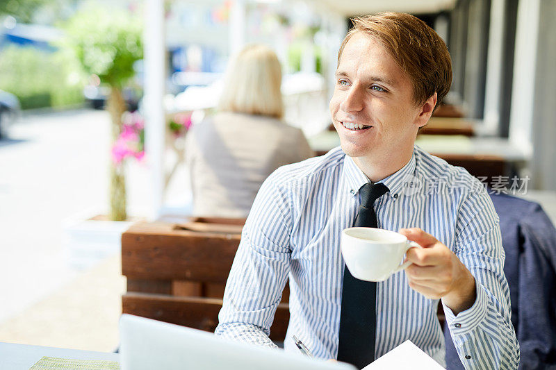 积极、有目标的年轻男性业务经理，穿着衬衫，坐在桌子旁喝咖啡，看着别处，在路边的咖啡馆
