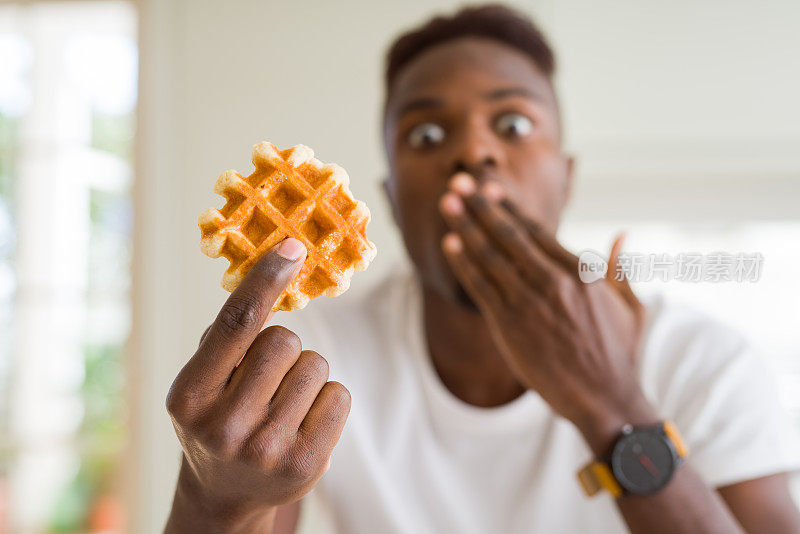 一名非裔美国男子吃着甜的比利时华夫饼捂着嘴用手震惊，为错误而羞愧，表情恐惧，默默害怕，秘密概念