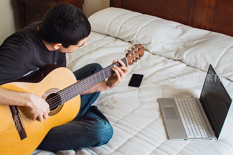 年轻人在床上弹吉他，用电脑在家学习。在家休闲:音乐和科技，通过在线课程学习吉他。