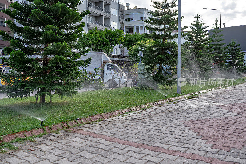 草坪灌溉在一个公园区域的散步在阿兰亚。水喷系统与城市景观和人行道的背景相映衬
