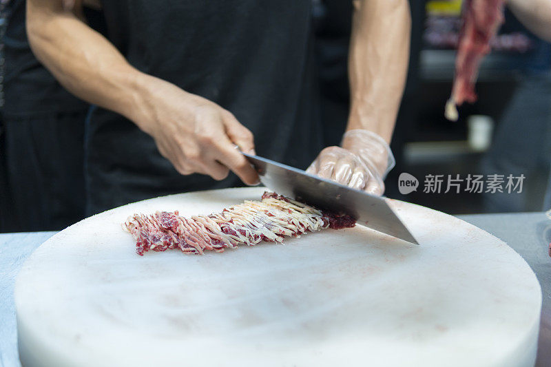 在一家牛肉火锅餐厅的开放式厨房里，厨师在挂着的切碎的生肉后面用刀切牛肉