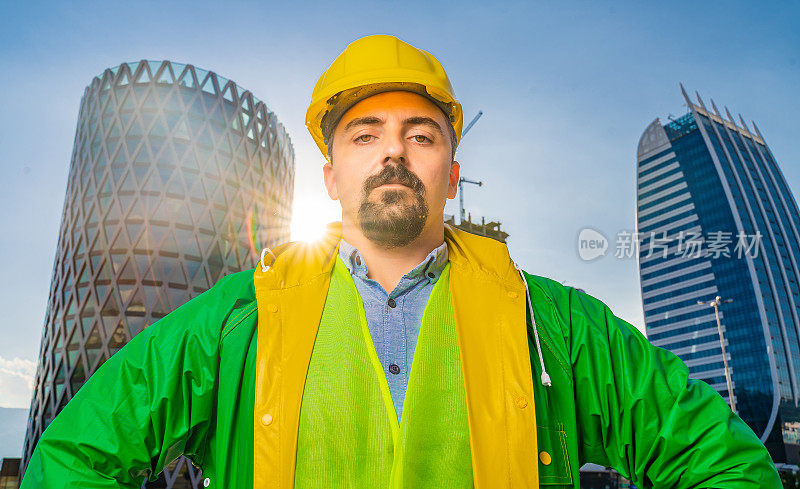 年轻工程师戴着黄色安全帽和绿色防护服在现代化的商业建筑和建筑工地前看着摄像机。镜头光晕。低角度的观点