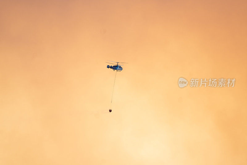 空中消防在黄色多云的天空背景中带着水篮的飞行直升机。