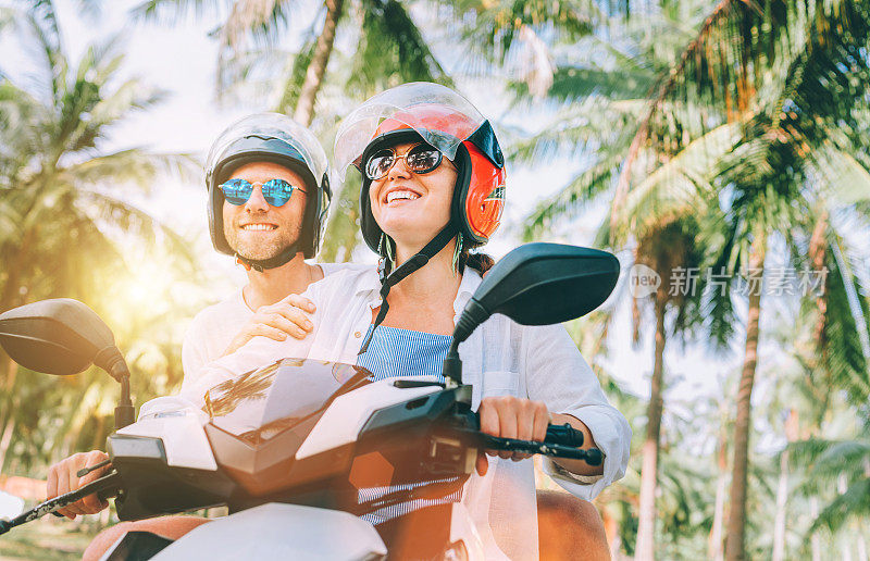 在泰国苏梅岛的棕榈树下，戴着安全帽骑着摩托车、面带微笑的夫妇在热带度假