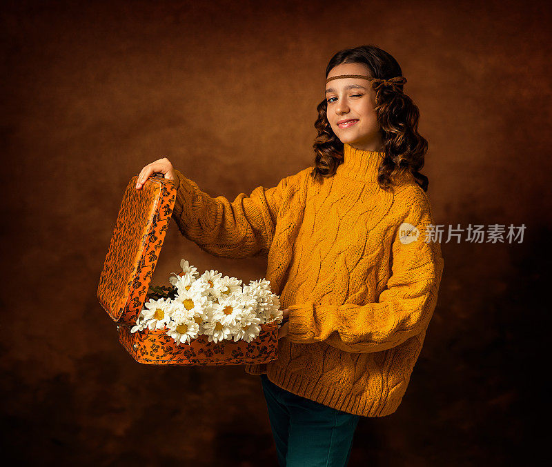 这个女孩手里拿着一个橘黄色的旧手提箱。她头上戴着一条缎带。手提箱里放着一束白色的菊花，雏菊。女孩眨眨眼，闭上一只眼睛。照片在复古风格。
