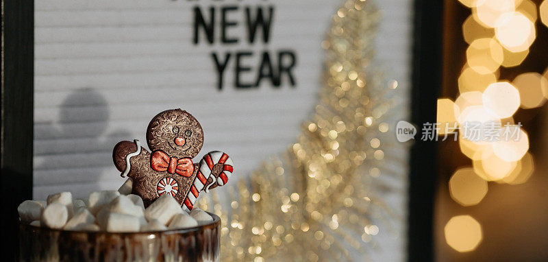 一个写着“新年快乐”的牌子，一个装有热可可、棉花糖的马克杯，一个姜饼人以美丽的黄色焦景为背景。一个温馨的圣诞和寒假概念。