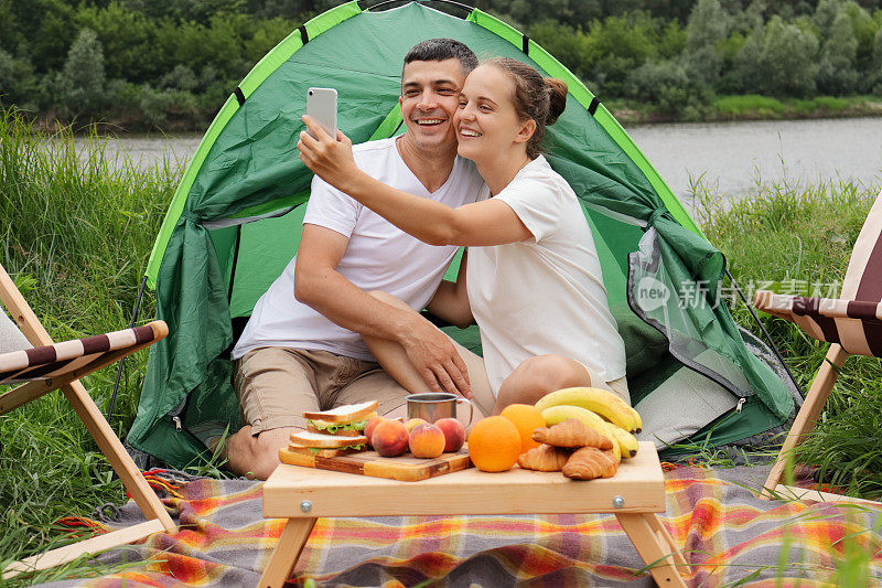 快乐的女人和男人穿着休闲的衣服坐在河边的帐篷里，用智能手机自拍，享受在大自然中休息。