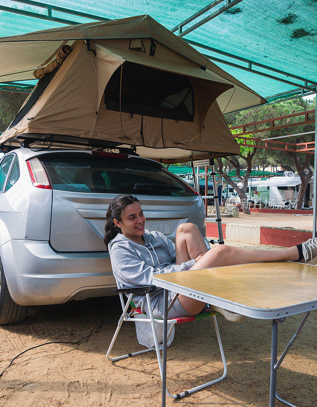 在一个露营地，一名年轻女子坐在椅子上看她的智能手机，她坐在车顶上搭着帐篷的汽车旁边。