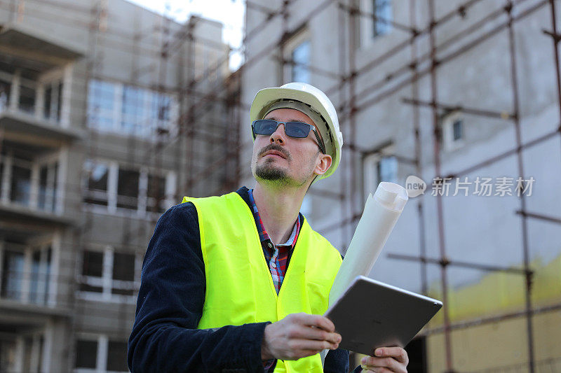 男建筑师在工地使用数码平板电脑，他穿着防护背心和头盔