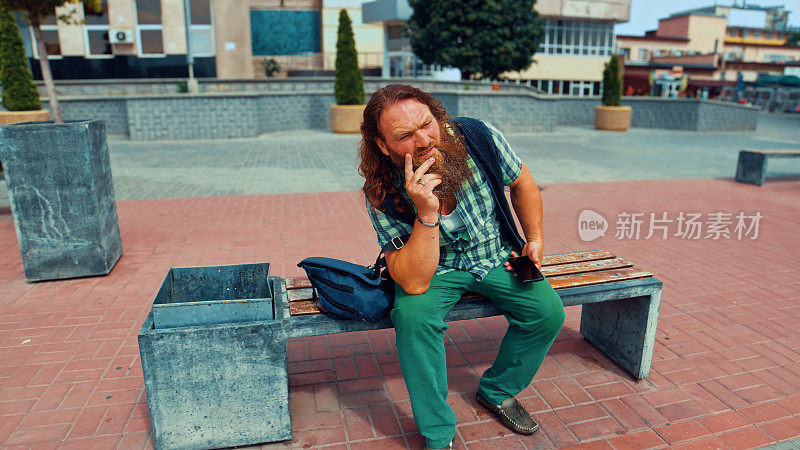 一位红发大胡子的老人坐在铺着红色石板的广场上的长凳上，手里拿着电话